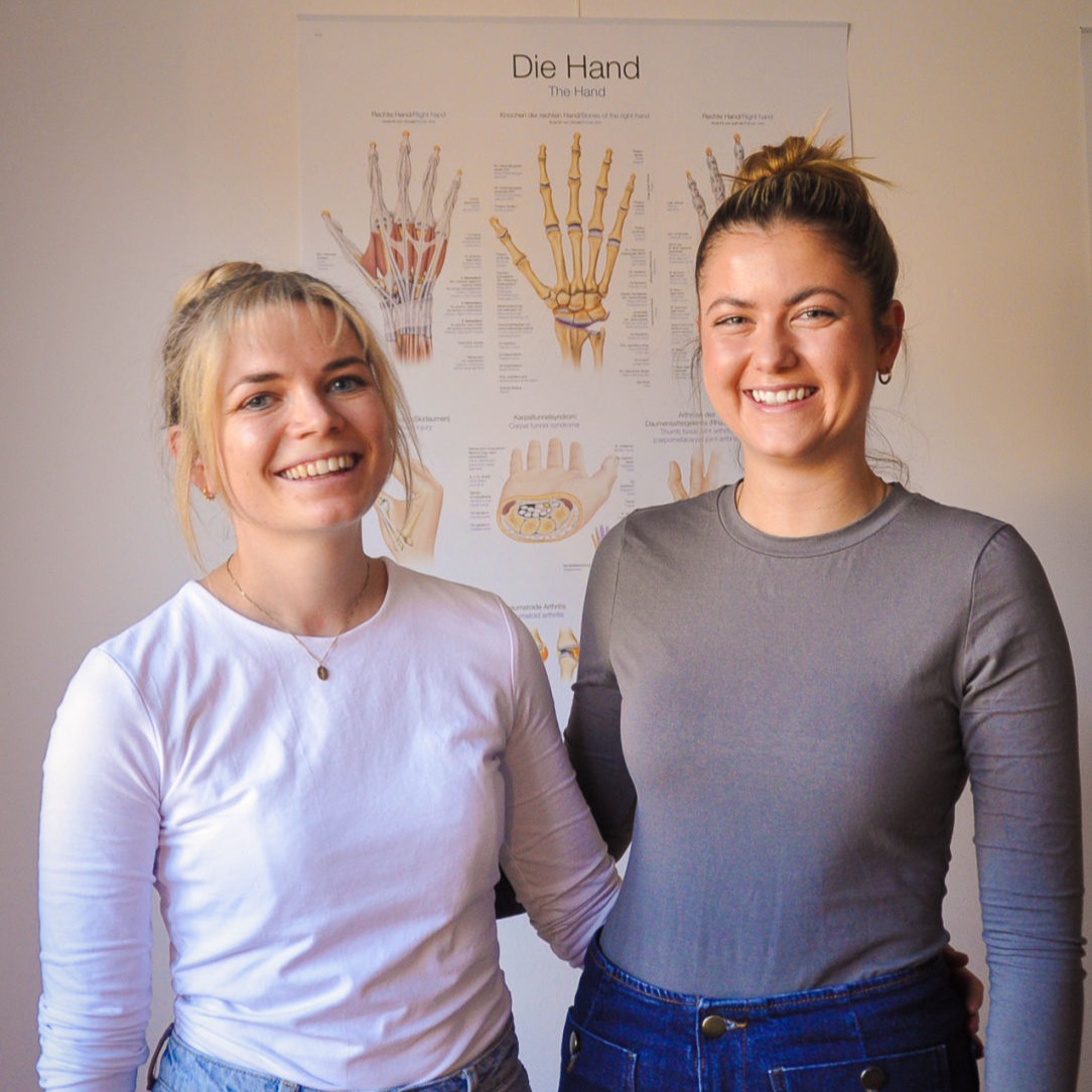 Lena Frank und ihr Kollegin in der Praxis vor medizinischem Poster zur Handtherapie